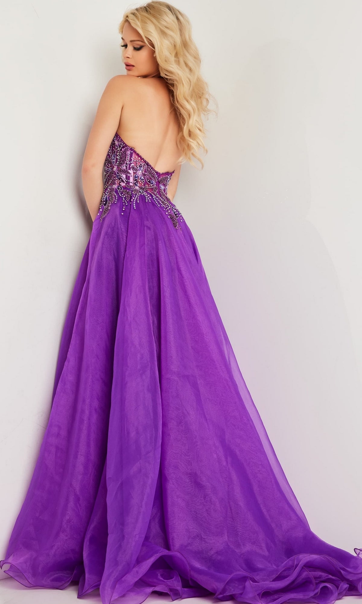 A woman with long blonde hair wears the Jovani Long Organza Halter Prom Ball Gown 25964, featuring beaded embellishments, as she stands against a light-colored wall.